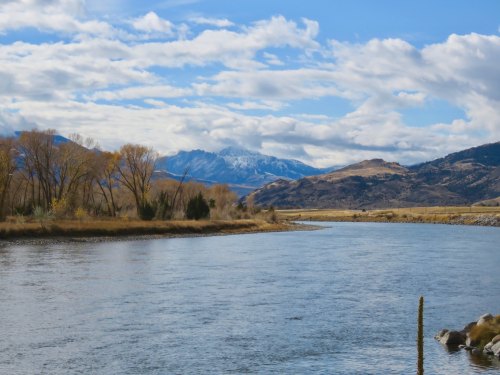 A serene river flows through a landscape of trees and mountains under a partly cloudy sky.