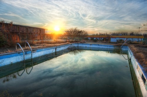 Abandoned swimming pool at sunset, surrounded by overgrown grass and a dilapidated building in the background.