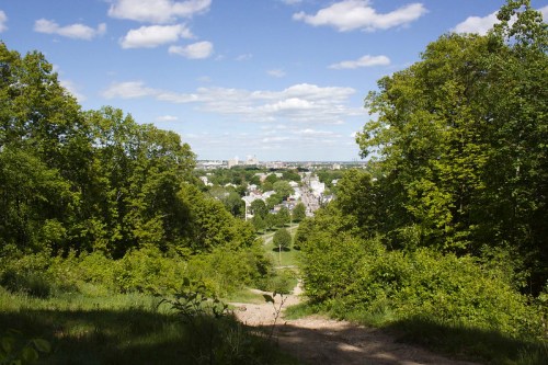 A scenic view from a hilltop overlooking a town, surrounded by lush greenery and a blue sky with fluffy clouds.