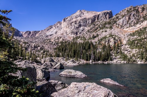 A serene mountain landscape featuring a clear lake, rocky shore, and towering peaks under a blue sky.