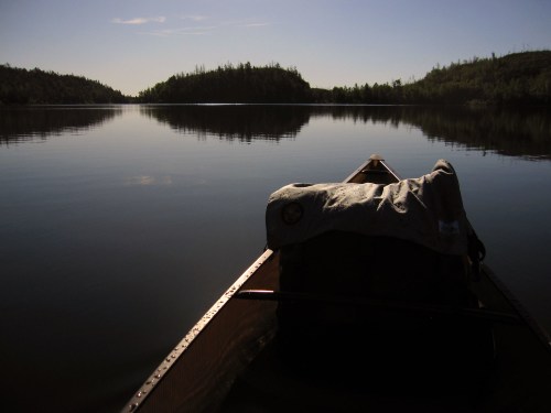 A canoe on calm water, with a blanket draped over the back, reflecting trees and a serene landscape in the distance.