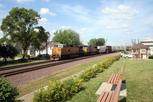 A train passes through a small town, with green grass and benches in the foreground under a blue sky.