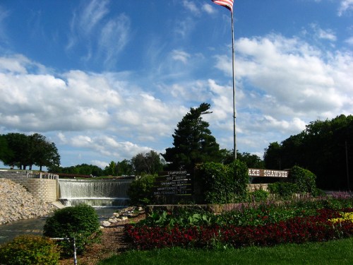 A scenic view of Lake Shawnee with a waterfall, colorful flowers, and an American flag against a blue sky.