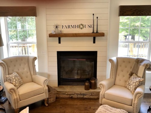 Cozy living room with two armchairs, a fireplace, and a farmhouse sign above a wooden shelf. Natural light from windows.