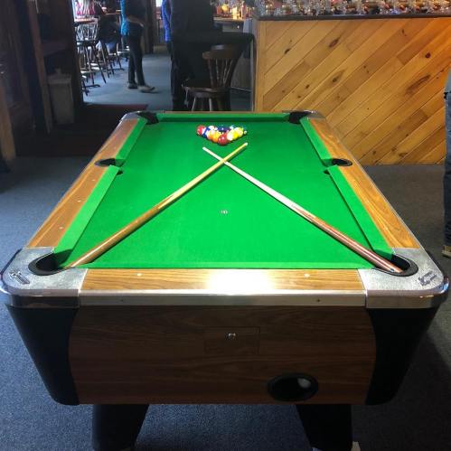 A pool table with a green felt surface, two cues crossed over a triangle of colored balls, set in a casual bar environment.