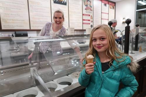A girl in a teal jacket smiles with an ice cream cone, while a server grins behind the ice cream counter.