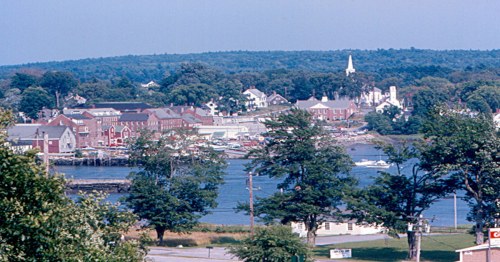 A scenic view of a coastal town with boats, trees, and buildings along the water, set against a green hillside.