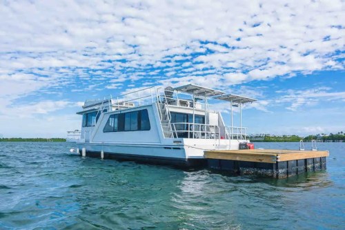 A white houseboat docked on calm water under a blue sky with scattered clouds.
