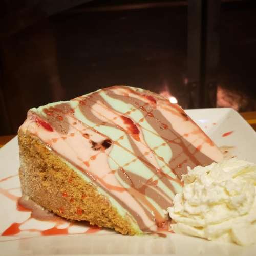 A slice of layered ice cream cake with chocolate and strawberry drizzle, served with a dollop of whipped cream.
