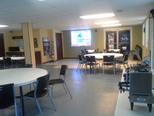 A spacious room with round tables, chairs, a projector screen, and a snack area, designed for gatherings or meetings.