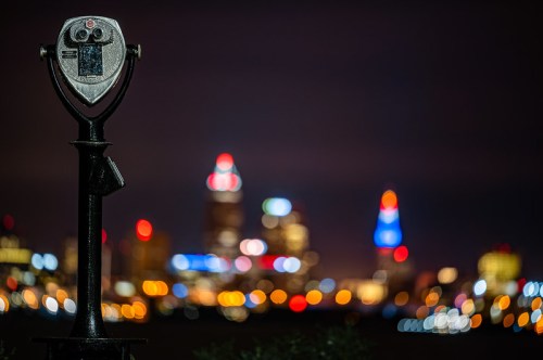 A coin-operated viewing telescope in the foreground with a blurred city skyline illuminated at night in the background.