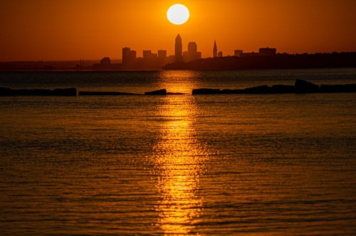 Sunset over a city skyline, with golden light reflecting on calm water and silhouettes of buildings in the background.
