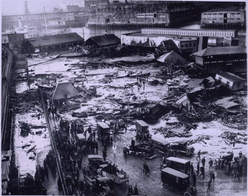 Aerial view of a disaster site with damaged buildings, debris, and people gathered, early 20th century.