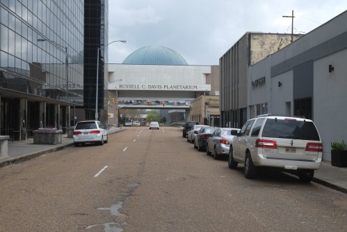 View of a street leading to the Russell C. Davis Planetarium, with parked cars and buildings on either side.