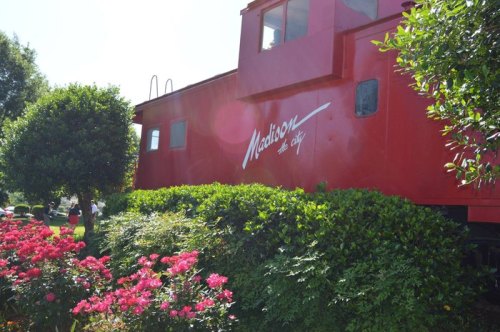 A bright red train caboose surrounded by green bushes and pink flowers in a sunny outdoor setting.