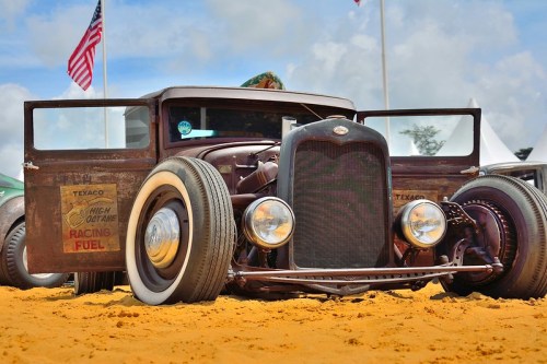 A vintage hot rod with a rusty finish parked on sandy ground, featuring Texaco racing fuel signage and open doors.