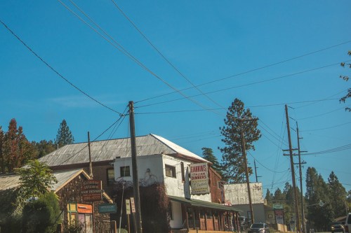 A small town scene featuring a vintage building with a sign, surrounded by power lines and tall trees under a clear blue sky.