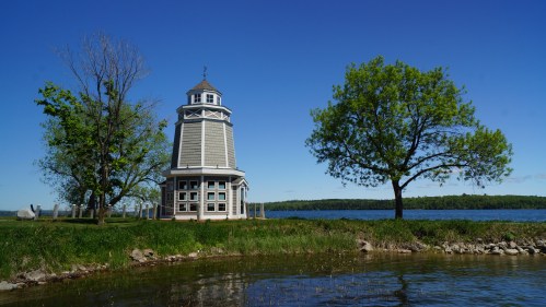 A tall, gray lighthouse beside a calm lake, surrounded by green trees and a clear blue sky.