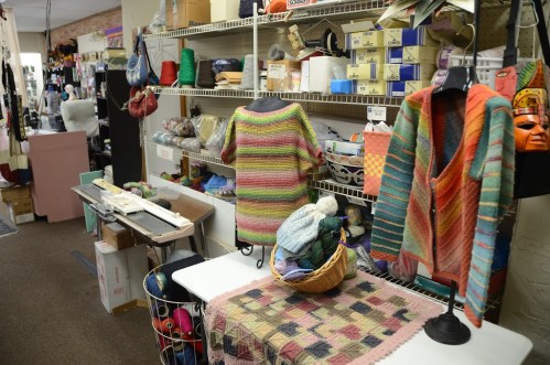 A cozy craft store interior with colorful yarn, knitted garments on display, and various crafting supplies on shelves.