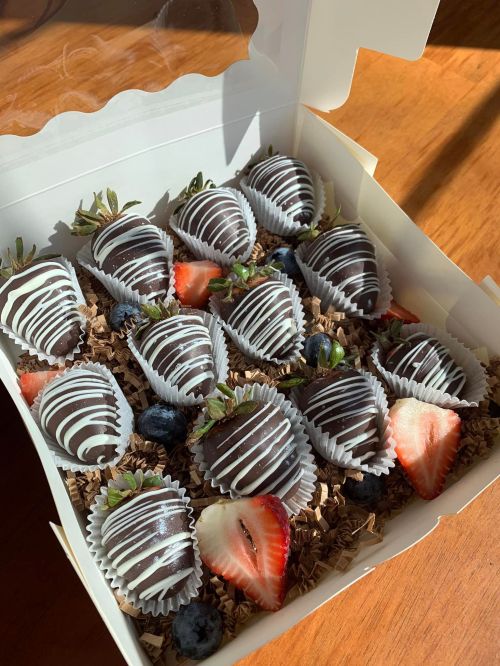 A box of chocolate-covered strawberries, drizzled with white chocolate, alongside fresh strawberries and blueberries.