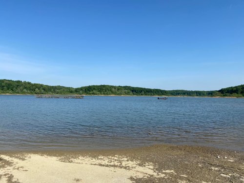 A serene lake scene with calm water, a sandy shore, and green hills under a clear blue sky.