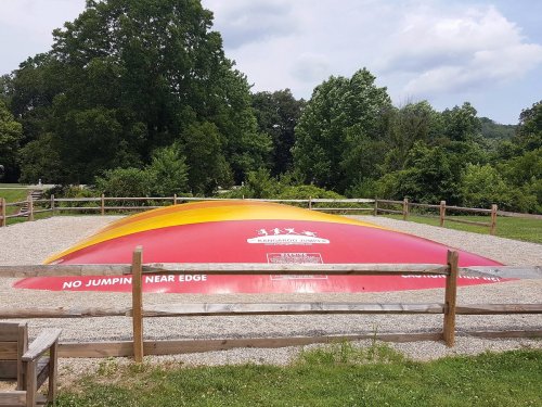 A large inflatable jumping pillow in a park, surrounded by a wooden fence and greenery under a cloudy sky.