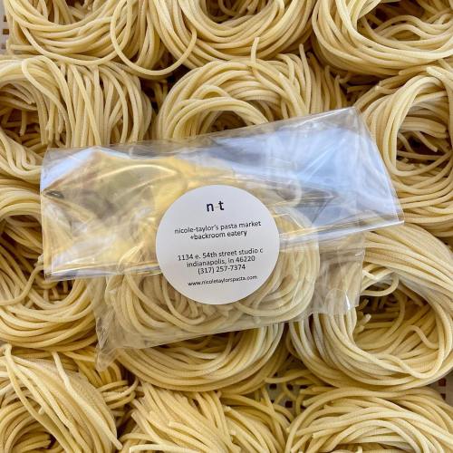 A clear bag of pasta from Nicole-Taylor's Pasta Market, surrounded by strands of pasta on a textured surface.