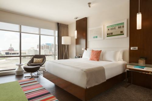 Modern hotel room with a large window, a queen bed, colorful rug, and stylish furniture, offering a city view.