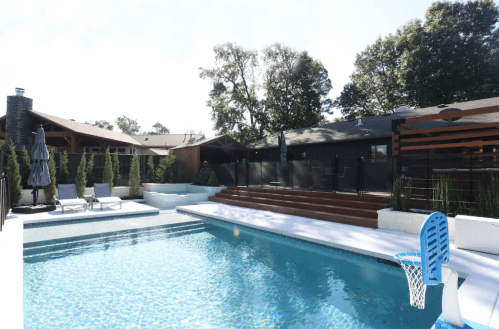 A serene backyard pool with lounge chairs, a basketball hoop, and surrounding greenery on a sunny day.
