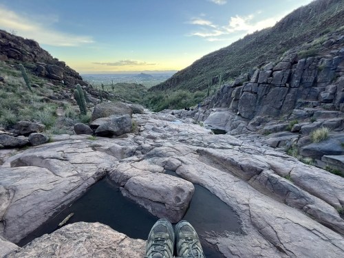A pair of hiking shoes on rocky terrain, overlooking a valley with distant mountains under a clear sky.