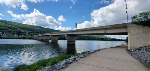 A concrete bridge spans a river, with green hills and fluffy clouds in the background, alongside a paved walkway.