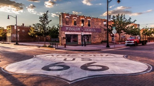 Historic Route 66 sign on a street in Winslow, Arizona, with vintage buildings and a red truck in the background.