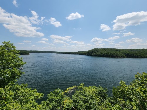 A serene lake surrounded by lush greenery under a bright blue sky with fluffy white clouds.