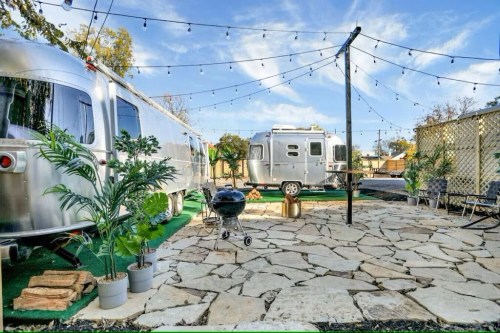 A cozy outdoor space featuring two silver trailers, string lights, a stone patio, and potted plants.