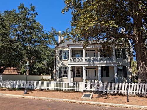 Historic two-story house with a white picket fence, surrounded by trees and a clear blue sky.