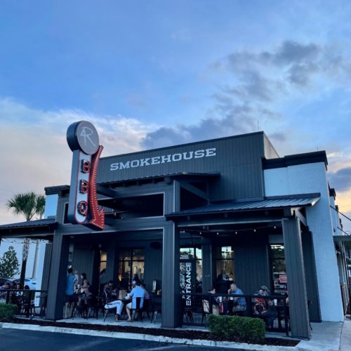 Exterior of a smokehouse restaurant with a retro sign, outdoor seating, and a cloudy sky in the background.