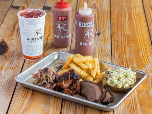 A tray with pulled pork, brisket, fries, coleslaw, and BBQ sauces, alongside a drink on a wooden table.
