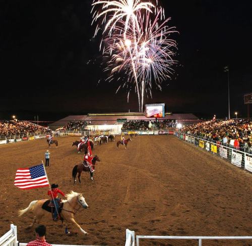 A rodeo event at night with riders on horseback, one holding an American flag, and fireworks lighting up the sky.