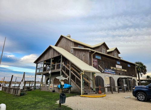 A wooden building with a sloped roof, featuring stairs and a sign for "Up the Creek Bar," near a waterfront.