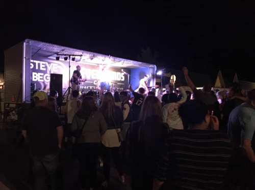 A crowd enjoys a live band performance at night, with a stage and colorful lights in the background.