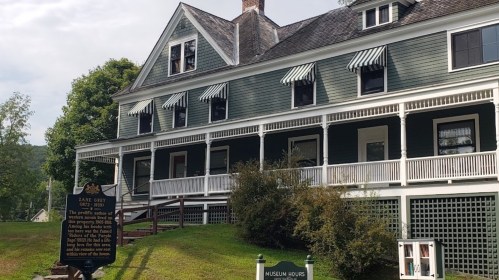 Historic house with green siding, white trim, and striped awnings, featuring a sign and museum hours displayed in front.