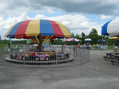A colorful carousel with a large umbrella, surrounded by empty picnic tables and amusement park attractions under a cloudy sky.