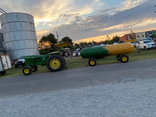 A green tractor pulls a corn-shaped trailer near a silos at sunset, with people and cars in the background.