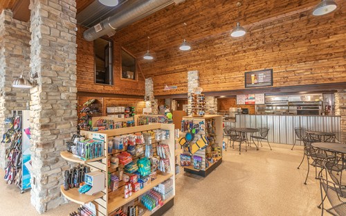 Interior of a rustic store with wooden beams, stone pillars, shelves of merchandise, and seating areas.