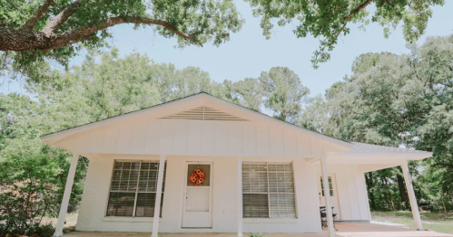 A white house with a front porch, surrounded by trees, featuring a decorative wreath on the door.