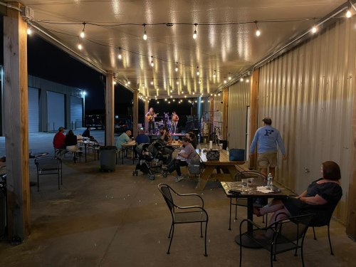 Outdoor gathering under string lights, with people seated at tables enjoying live music performance in the background.