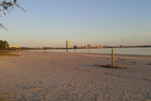 A serene beach at sunset, with calm water reflecting buildings in the distance and a clear sky.