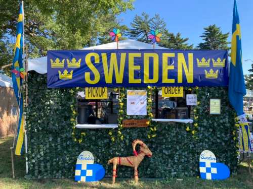 A festive booth decorated with greenery and flags, featuring "SWEDEN" signage and a wooden goat statue.