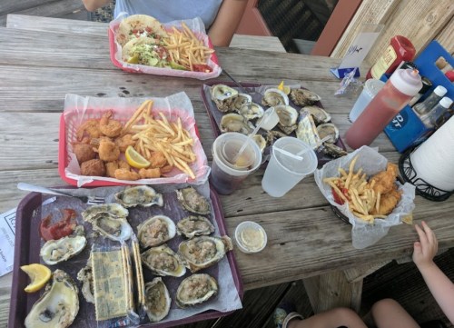 A table with trays of fried seafood, oysters, fries, and drinks, set outdoors on a wooden surface.