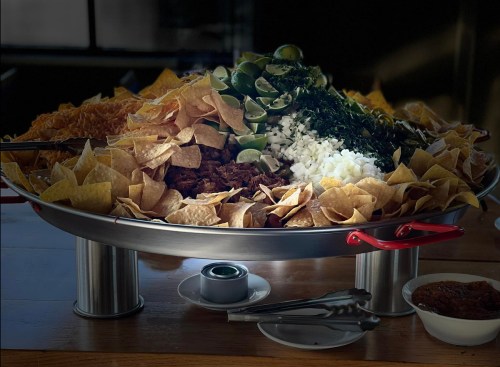 A large platter filled with tortilla chips, meat, onions, limes, and greens, served with small bowls of sauce.